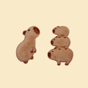 May include: Two tan-colored capybara plushies on a cream background. One capybara stands upright, looking up. The other is a stack of three capybaras. The capybaras have red-brown noses and ears, and small black eyes.