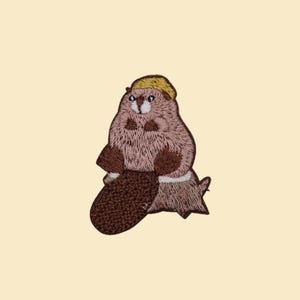 May include: Embroidered patch featuring a cartoon beaver wearing a yellow hard hat. The beaver is brown and tan with a large, dark brown tail. The patch is on a light beige background.