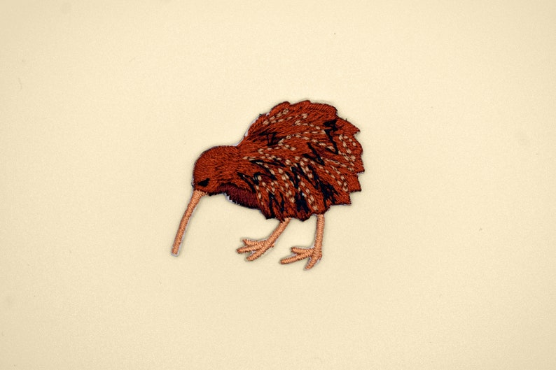 Op de afbeelding: Een bruin geborduurd pleister van een kiwi-vogel met een lange snavel en kleine poten. De vogel kijkt naar links en heeft een bruin en zwart verenontwerp.