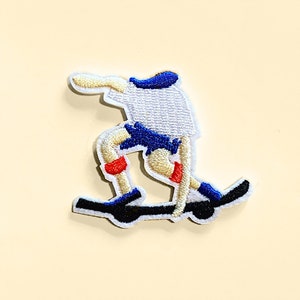 May include: Embroidered iron-on patch of a person skateboarding. The person is wearing a white shirt, blue shorts, and red and blue shoes. The skateboard is black.