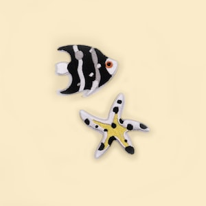 May include: Two embroidered patches: a black and white striped fish with an orange eye and a white starfish with black spots and a yellow center. The patches are on a light yellow background.