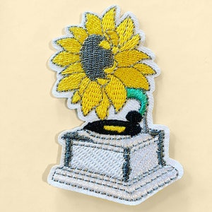 May include: A white embroidered patch of a yellow sunflower with a green stem on top of a white vintage record player.
