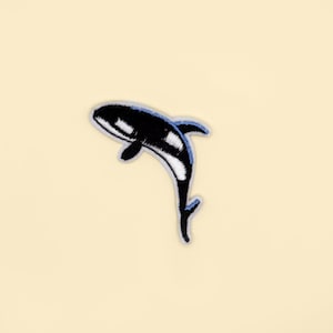 May include: Embroidered patch depicting a black and white orca whale with a blue border. The whale is shown leaping, set against a cream background. Ideal for customising clothing or accessories, this patch is a fun addition.