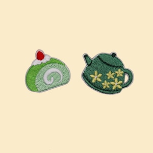 May include: Embroidered patches featuring a green cake roll with a red cherry and a green teapot decorated with yellow flowers. The patches are on a light yellow background. These patches are perfect for adding to clothing or accessories.