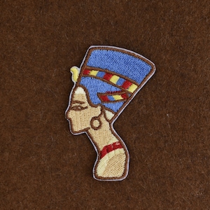 May include: Embroidered patch featuring the profile of an Egyptian queen, with a blue headdress and gold accents. The patch has a tan background and a white border. The design is detailed and colorful, suitable for clothing or accessories.