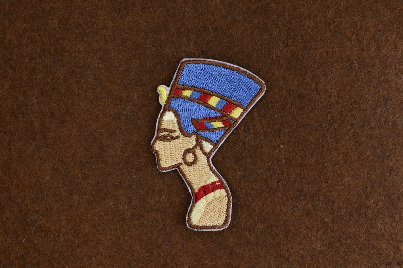 May include: Embroidered patch featuring the profile of an Egyptian queen, with a blue headdress and gold accents. The patch has a tan background and a white border. The design is detailed and colorful, suitable for clothing or accessories.