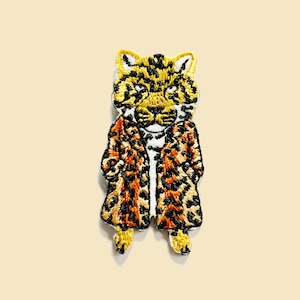 Op de afbeelding: Een geborduurde strijk-patch met een cartoon-luipaard dat een luipaardprintjas draagt. De luipaard heeft een geel-zwarte gevlekte vacht en een wit gezicht met zwarte ogen en een zwarte neus.