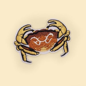 Op de afbeelding: Geborduurde krabpatch met een bruin lichaam en gele scharen. De krab heeft een wit geborduurd ontwerp op zijn lichaam. De patch staat op een lichtgele achtergrond.