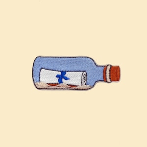 Puede incluir: Una botella de vidrio azul con un tapón de corcho contiene un papel blanco enrollado con una cinta azul. La botella está sobre una superficie de color beige.