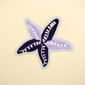 May include: Embroidered starfish patch in shades of purple and white, with a light grey border. The starfish design is centered on a cream-coloured background. Suitable for clothing or accessories.