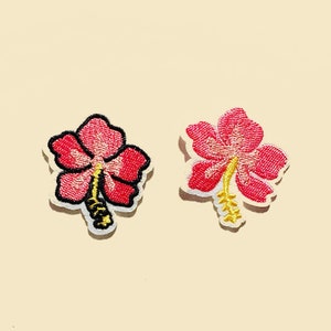 May include: Two red embroidered hibiscus flower patches with yellow centres and stems. The flowers are on a light beige background.