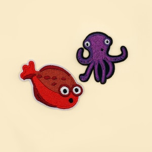 Könnte beinhalten: Zwei gestickte Aufnäher: ein roter Fisch mit weißen Augen und ein lila Oktopus mit schwarzen Umrissen. Der Fisch hat einen überraschten Gesichtsausdruck. Der Oktopus hat ein einfaches Design. Beide Aufnäher befinden sich auf einem cremefarbenen Hintergrund.