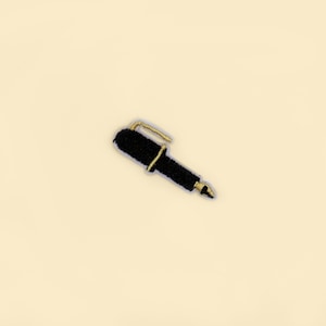 May include: A black pen with a gold-colored clip and tip. The pen is set against a cream-colored background. The pen appears to be a decorative item or an accessory.
