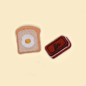 May include: Embroidered patches of breakfast foods on a light yellow background. One patch is a slice of toast with a fried egg, and the other is a rectangular piece of toast with the word "love" embroidered on it.