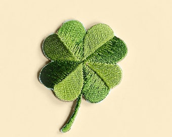 May include: Embroidered four-leaf clover patch in shades of green. The clover has four distinct leaves with detailed stitching, and a small stem. The patch is set against a light beige background, creating a contrast that highlights the clover's design.