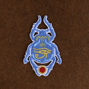 May include: Embroidered patch featuring a blue scarab beetle design with yellow and red accents. The beetle has an eye symbol and other hieroglyphic details. The patch is likely for clothing or accessories.
