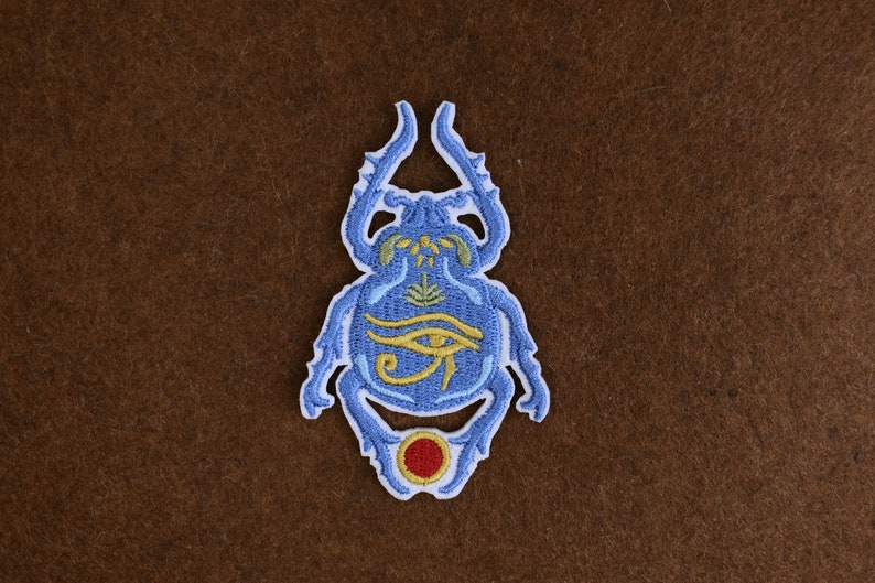 May include: Embroidered patch featuring a blue scarab beetle design with yellow and red accents. The beetle has an eye symbol and other hieroglyphic details. The patch is likely for clothing or accessories.