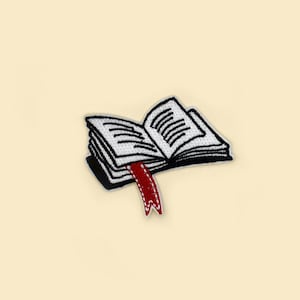 Op de afbeelding: Een geborduurde patch in zwart-wit van een open boek met een rode bladwijzer. Het boek is open op een pagina met regels tekst.