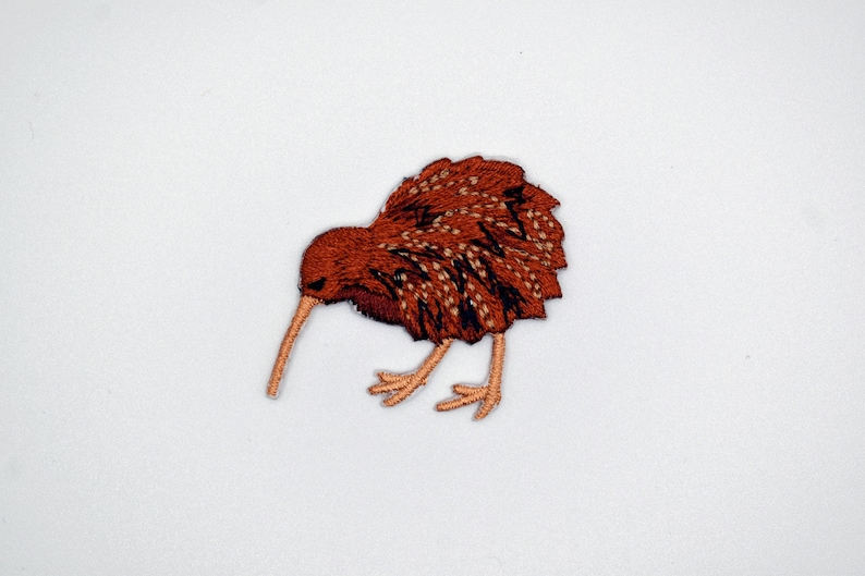 Op de afbeelding: Bruin geborduurd kiwi-vogelpatch met een lange snavel en kleine poten. De vogel kijkt naar links en heeft een bruin en zwart verenontwerp.