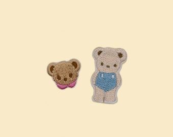 May include: Two embroidered iron-on patches featuring brown teddy bears. One bear is wearing blue shorts.