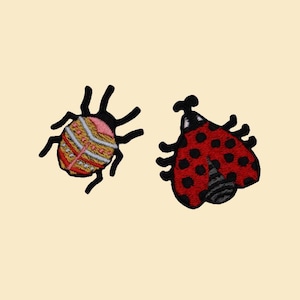 Op de afbeelding: Twee geborduurde insectenpatches. Eén heeft een kleurrijk gestreept ontwerp met zwarte poten en antennes. De andere is een rode lieveheersbeestje met zwarte stippen en antennes. Beide op een crèmekleurige achtergrond.
