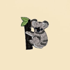 May include: Embroidered iron-on patch featuring a gray koala bear sitting on a black tree branch with green leaves.