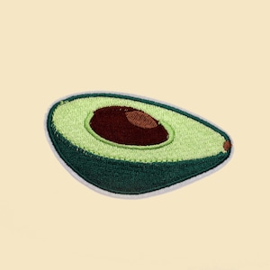 Op de afbeelding: Een groene geborduurde avocadohelft met een bruine pit, op een lichtgele achtergrond.