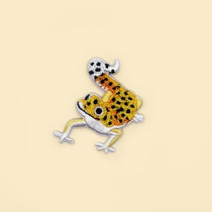 Pode incluir: Um remendo de geco bordado em amarelo e preto com costuras prateadas. O geco está virado para a direita e tem a cauda enroscada.