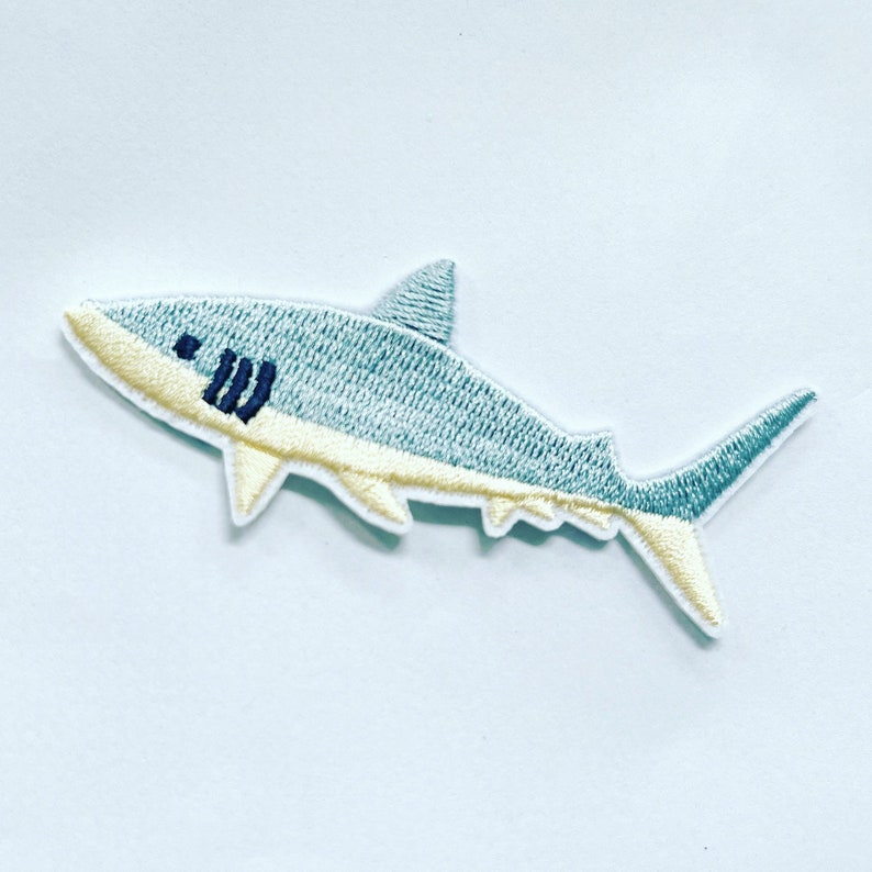 May include: A blue and yellow embroidered iron-on patch of a shark. The shark is facing left and has a white outline.