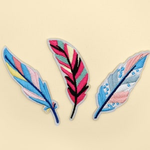 May include: Three colourful embroidered fabric patches in the shape of feathers. The feathers are in shades of blue, pink and yellow.