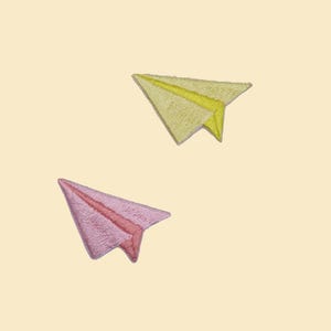 May include: Two embroidered paper airplane patches. One is yellow with a yellow accent, and the other is pink with a red accent. The patches are on a light beige background. These patches can be used to decorate clothing or accessories.