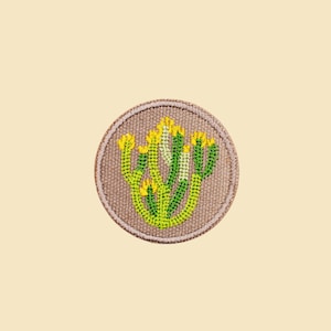 Op de afbeelding: Een ronde, beige stoffen patch met een geborduurd ontwerp van een groene en gele cactusplant.