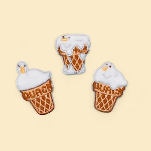 Puede incluir: Tres parches bordados con un diseño de cono de helado con forma de pato. El helado es blanco, el cono es marrón y la palabra "QUACK" está bordada en el cono. Los parches están sobre un fondo amarillo claro.