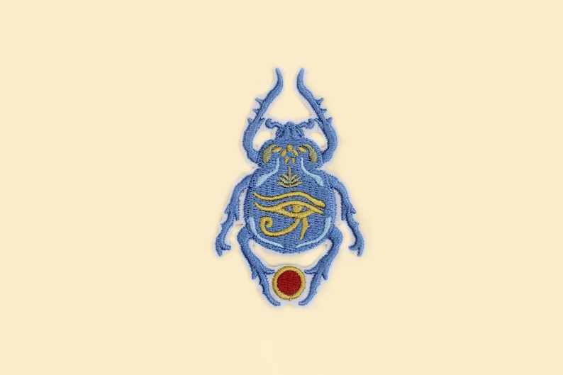 May include: Embroidered patch featuring a blue scarab beetle design with gold accents and an Eye of Horus symbol. The patch has a red circle at the bottom and is set against a cream background. This patch is suitable for clothing or accessories.