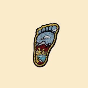 May include: A yellow and black embroidered patch of a footprint with a landscape scene inside. The scene includes mountains, trees, a lake, and clouds.