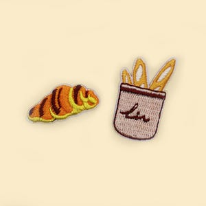 Könnte beinhalten: Zwei gestickte Bügelbilder, eines mit einem Croissant und das andere mit einer Tüte Brot mit dem Wort "Lin" darauf.