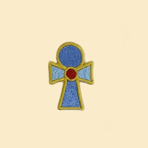 May include: Embroidered patch featuring an Ankh symbol, a cross with a loop at the top. The patch has a blue center, a red dot, and gold and light blue trim. The background is a light beige.