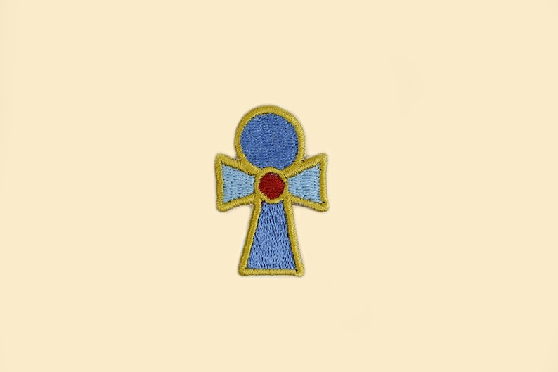 May include: Embroidered patch featuring an Ankh symbol, a cross with a loop at the top. The patch has a blue center, a red dot, and gold and light blue trim. The background is a light beige.