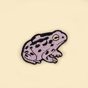 Könnte beinhalten: Gestickter Aufnäher mit einer stilisierten Froschdarstellung in Lavendel und Schwarz. Der Frosch hat ein detailliertes Design mit schwarzer Umrandung und schwarzen Punkten. Der Aufnäher befindet sich auf einem hellbeigen Hintergrund.