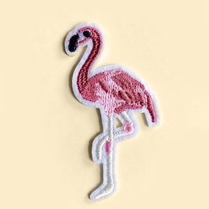May include: Pink flamingo embroidered iron-on patch. The flamingo is standing on one leg with its head bent down. The patch is made of felt and has a white outline.
