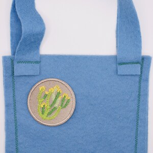 May include: A small blue felt tote bag with a green stitched border and a beige embroidered patch featuring a green plant with yellow flowers.