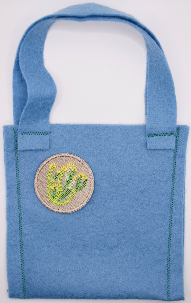 May include: A small blue felt tote bag with a green stitched border and a beige embroidered patch featuring a green plant with yellow flowers.