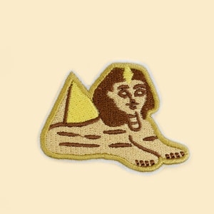 May include: Embroidered patch featuring a stylized Sphinx with a pyramid. The patch has a tan background with brown and yellow details. The Sphinx is depicted in a seated position, with a human face and a lion's body.