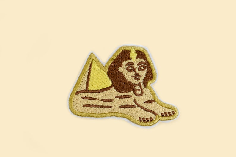 May include: Embroidered patch featuring a stylized Sphinx with a pyramid. The patch has a tan background with brown and yellow details. The Sphinx is depicted in a seated position, with a human face and a lion's body.