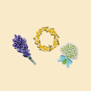 May include: Three embroidered floral designs on a pale yellow backdrop. A bundle of purple lavender, a yellow and white floral wreath, and a small bouquet of white flowers with a blue ribbon.
