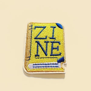 May include: Yellow embroidered iron-on patch with the word "ZINE" in blue thread. The patch is shaped like a book with a blue and white spine.