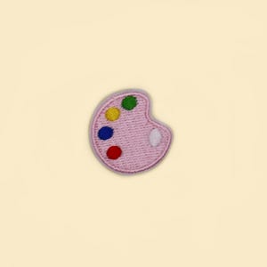 Puede incluir: Un parche bordado rosa con forma de paleta de pintor. La paleta presenta manchas de pintura coloridas en rojo, azul, amarillo, verde y blanco. El parche es un suministro de manualidades para los amantes del arte.
