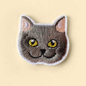 Op de afbeelding: Geborduurde strijkpatch van een grijs kattengezicht met gele ogen. De kat glimlacht.
