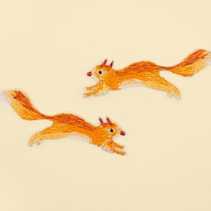 Peut inclure: Deux patchs de  écureuil brodés orange et blanc sur un fond jaune clair. Les écureuils sont tournés vers la droite et ont la queue enroulée.