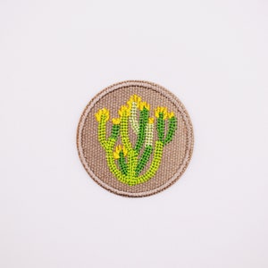 May include: A round, beige fabric patch with an embroidered cactus design in green and yellow.